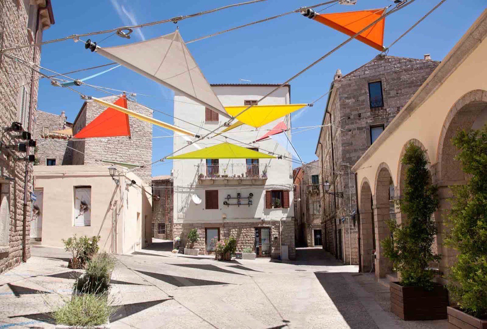 A quiet stone-paved street in Tempio Pausania, shaded by colourful triangular sails strung between the granite buildings